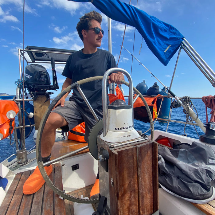 Man vid ratten på en segelbåt med orange Fubuki Nozawa-tofflor på fötterna